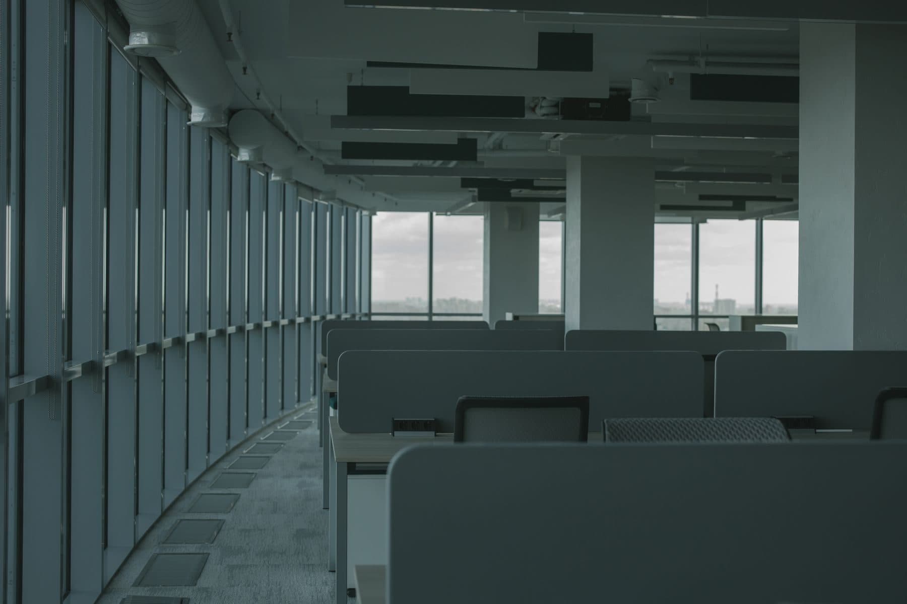 An empty open-plan office with long rows of desks and a wall of tall windows.